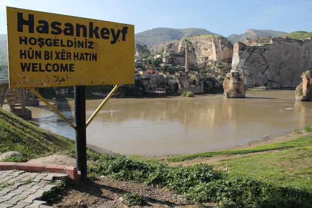 A road sign reading 'Hasankeyf — welcome' in Turkish, Kurdish, English and Arabic stands in the foreground; behind it the old town climbs a limestone cliff cut with cave openings, above the broad green Tigris. Photographed before the Ilısu reservoir filled.